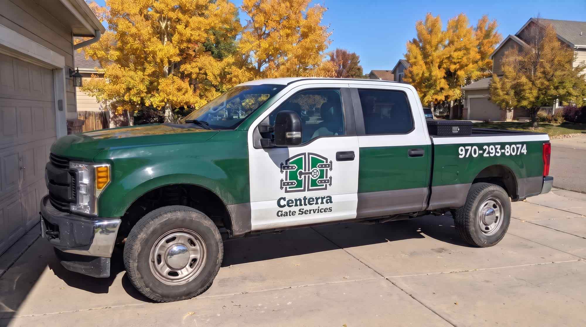 Green and white Centerra Gate Services crew-cab pickup in a driveway, the gate logo on the front door and 970-293-8074 on the bed, golden trees behind