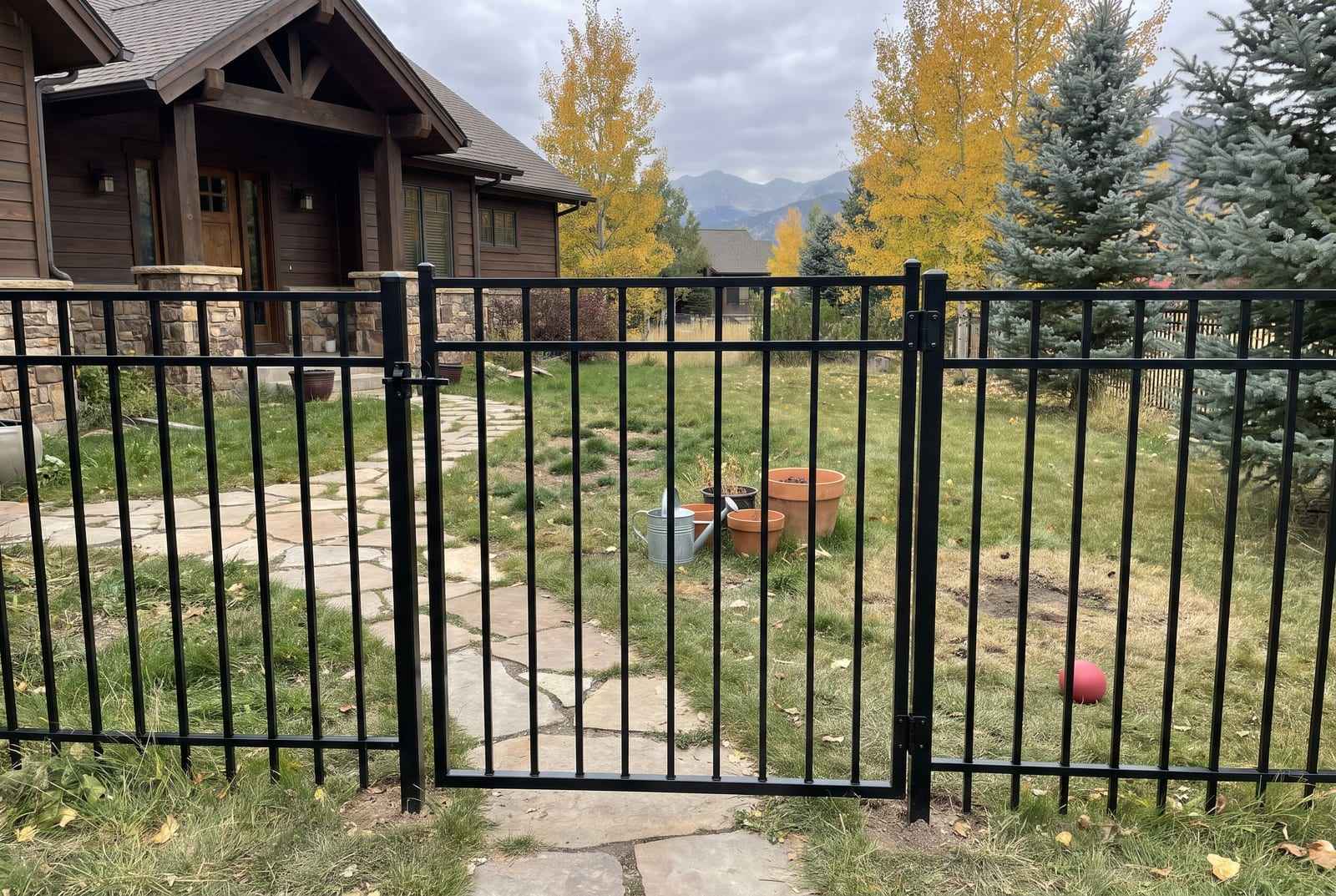 Black wrought-iron walk gate in a matching fence beside a flagstone path, golden trees and mountains behind