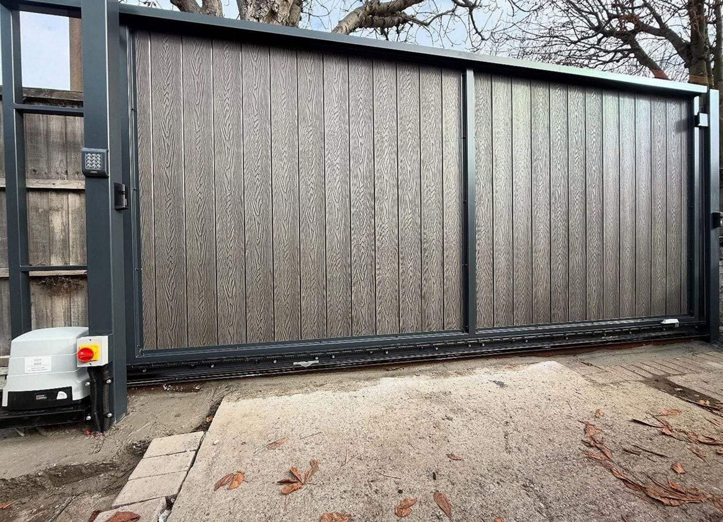 Grey woodgrain composite sliding gate in a dark steel frame, its opener and isolator switch at the post and a keypad on the far pillar