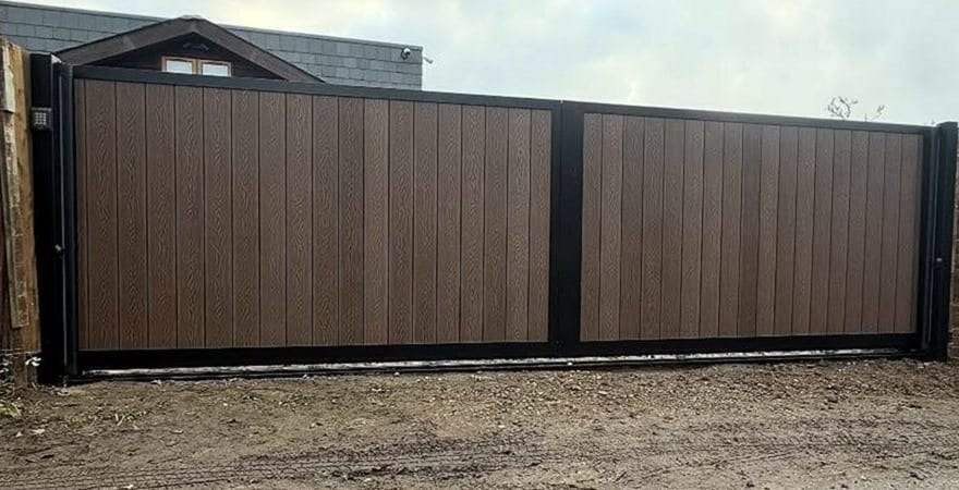 Wide brown composite sliding gate with two framed panels across a dirt drive, a keypad on the post at its left