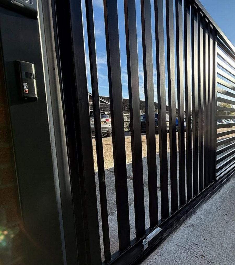 Tall black vertical-bar sliding gate on a commercial lot, a sensor housing mounted on the steel post beside it