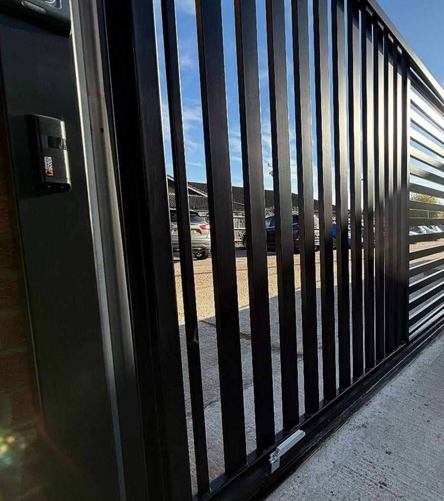 Tall black vertical-bar sliding gate on a commercial lot, a sensor housing mounted on the steel post beside it