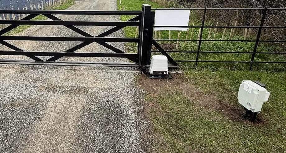 Black five-bar sliding gate across a gravel lane, its white opener housing at the post and a control box on a stand in the grass