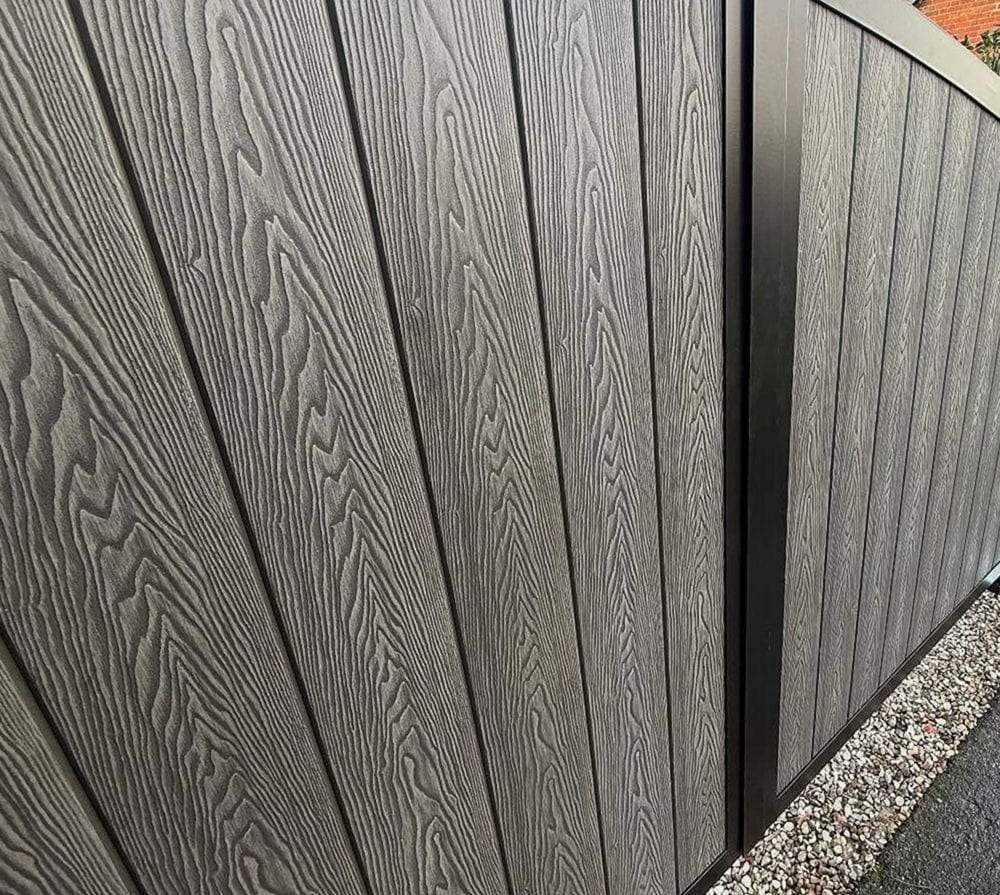 Close view of grey woodgrain composite swing gate panels in a black frame, the hinge post at the far end and gravel at its foot