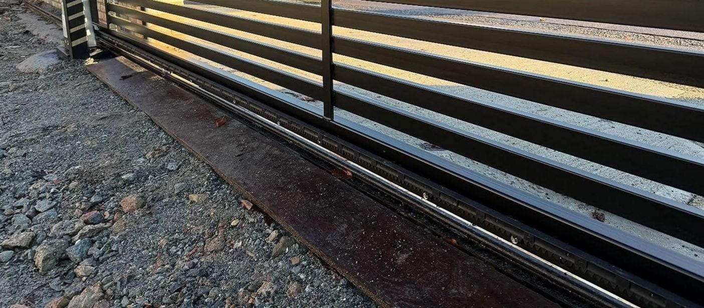 Bottom rail of a slatted sliding gate with its toothed gear rack and ground track running along a gravel drive