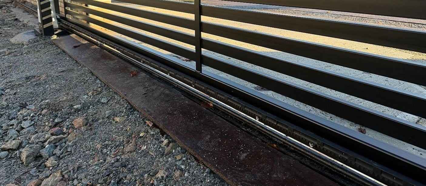 Bottom rail of a slatted sliding gate with its toothed gear rack and ground track running along a gravel drive